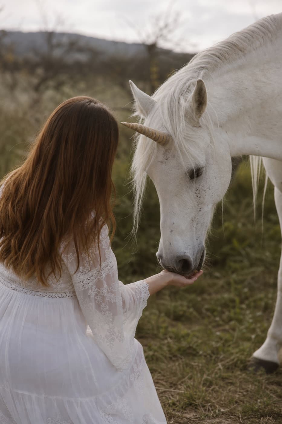A woman gently feeding a unicorn in a field, back to yourself